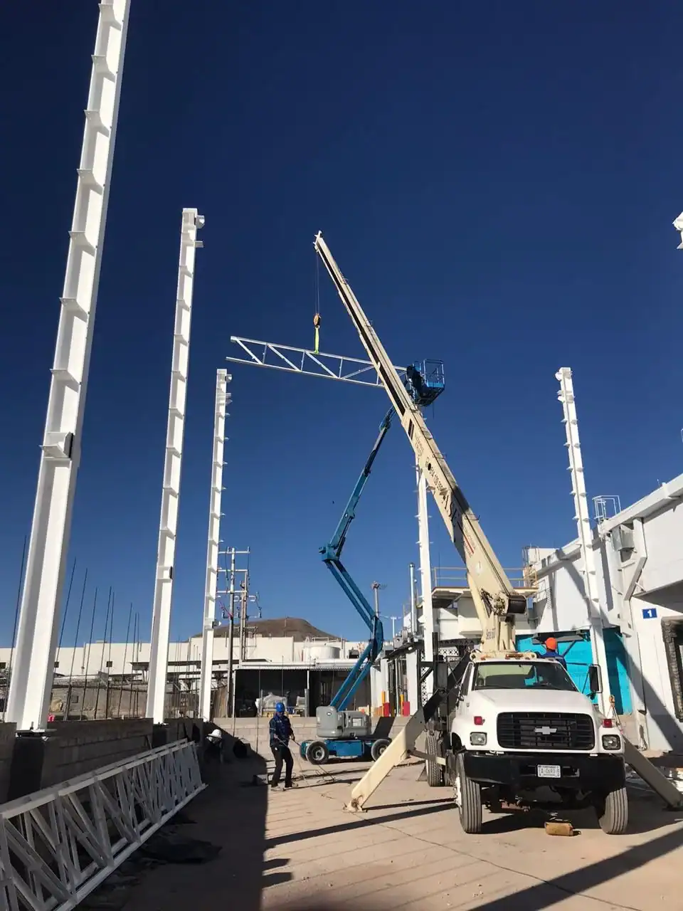 Obra de construcción con grúa elevando estructura metálica y operarios trabajando.