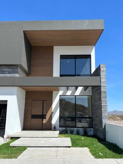 Fachada frontal de casa moderna de dos pisos con volúmenes en gris, blanco y madera, y entrada con jardín.